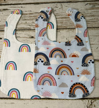 Load image into Gallery viewer, Two fabric bibs laid flat on a rustic whitewashed wooden surface. One bib is white with colorful rainbow prints, and the other is light blue with a matching rainbow and cloud pattern.
