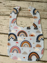 Load image into Gallery viewer, A light blue fabric baby bib with a colorful rainbow, sun, and cloud pattern, laid flat on a rustic light wood background.
