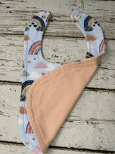 Load image into Gallery viewer, A reusable baby bib with a light blue and white rainbow pattern on one side and a solid peach-colored fabric on the other, displayed on a rustic white wooden surface.
