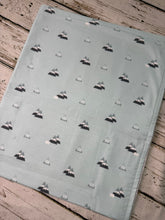 Load image into Gallery viewer, A baby blanket or swaddle, featuring a repeating pattern of small gray and black mountain triangles on a pale sage green background, laid flat on a rustic light wood plank surface.
