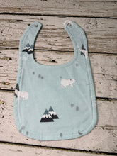 Load image into Gallery viewer, A light blue fabric baby bib with a printed pattern of polar bears, mountains, and pine trees, laid flat on a rustic wooden plank surface.
