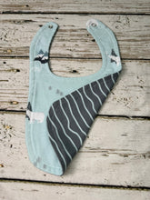 Load image into Gallery viewer, A baby bib with a light blue background, featuring a dark gray and white striped pattern on one side, and small white animal and mountain prints on the other side. The bib is laid flat on a rustic, weathered wood plank surface.
