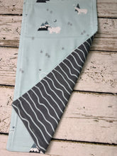 Load image into Gallery viewer, A folded baby burp cloth made of light blue fabric with a cute sheep and mountain print, showing a dark gray reverse side with white wavy stripes, laid flat on a rustic white-washed wooden surface.
