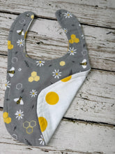 Load image into Gallery viewer, A baby bib with a gray and white honeybee and daisy pattern, laid flat on a rustic white-washed wooden surface. The bib features a white reverse side with yellow polka dots.
