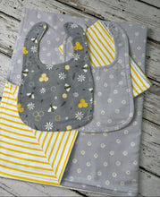 Load image into Gallery viewer, A baby gift set laid flat on a rustic white wood background. The set includes a blanket,  gray bib with a floral and bee pattern, a gray bib with white daisy and bee patterns, and yellow-and-white striped burp cloth.
