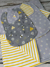 Load image into Gallery viewer, A baby gift set laid flat on a rustic white wood background. The set includes a blanket,  gray bib with a floral and bee pattern, a gray bib with white daisy and bee patterns, and yellow-and-white striped burp cloth.
