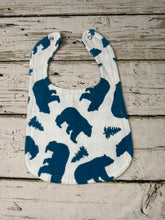 Load image into Gallery viewer, A white fabric baby bib with a dark blue pattern of bears and pine trees, laid flat on a rustic, light-colored wooden plank surface.
