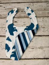 Load image into Gallery viewer, A baby bib with a white background, featuring dark blue bear silhouettes on one side and blue, navy, and white stripes on the other, laid flat on a rustic light wood plank surface.
